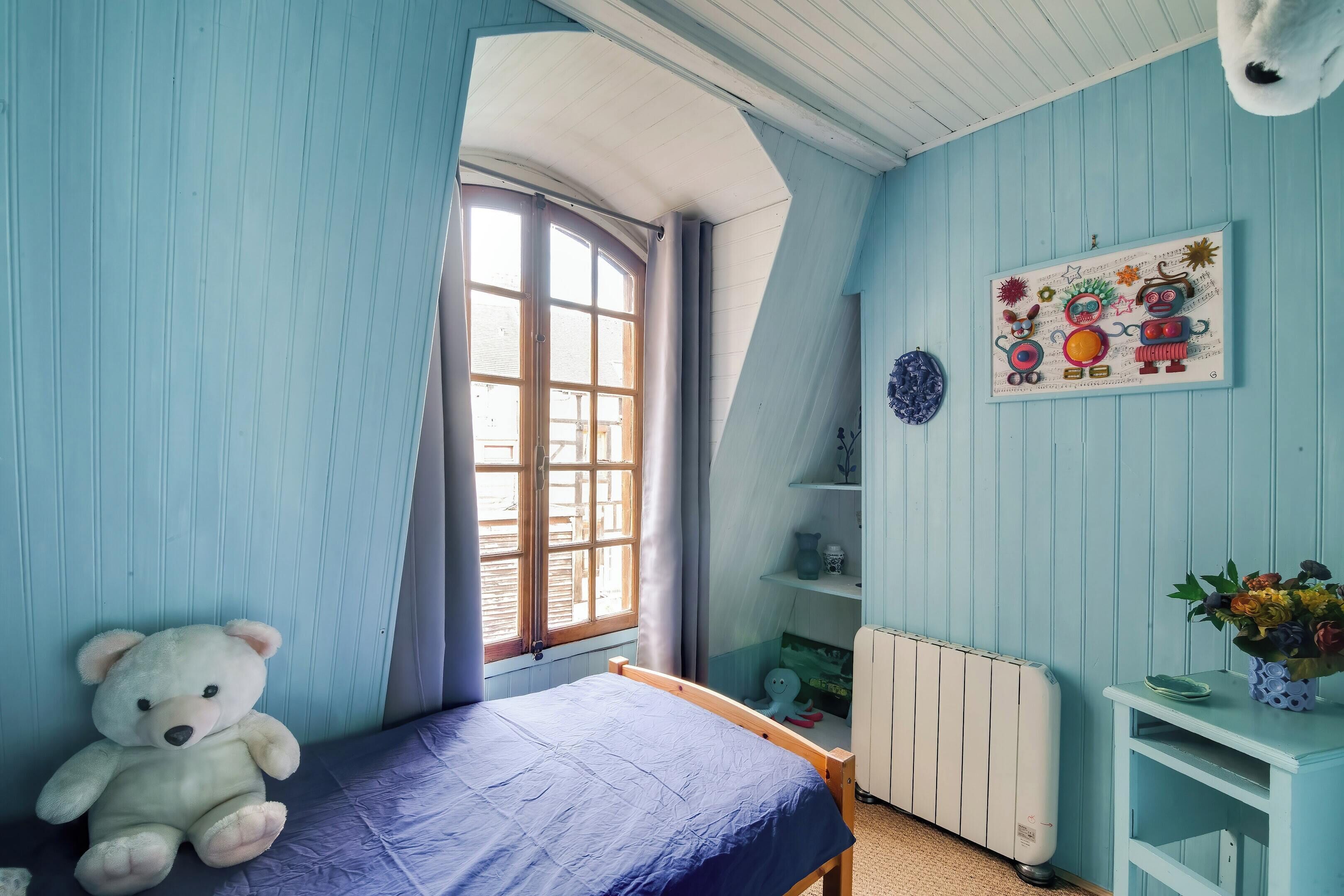 Une pièce avec un lit, un ours en peluche, une fenêtre, un radiateur et un mur orné d’un tableau coloré.