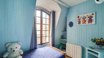 Une pièce avec un lit, un ours en peluche, une fenêtre, un radiateur et un mur orné d’un tableau coloré.