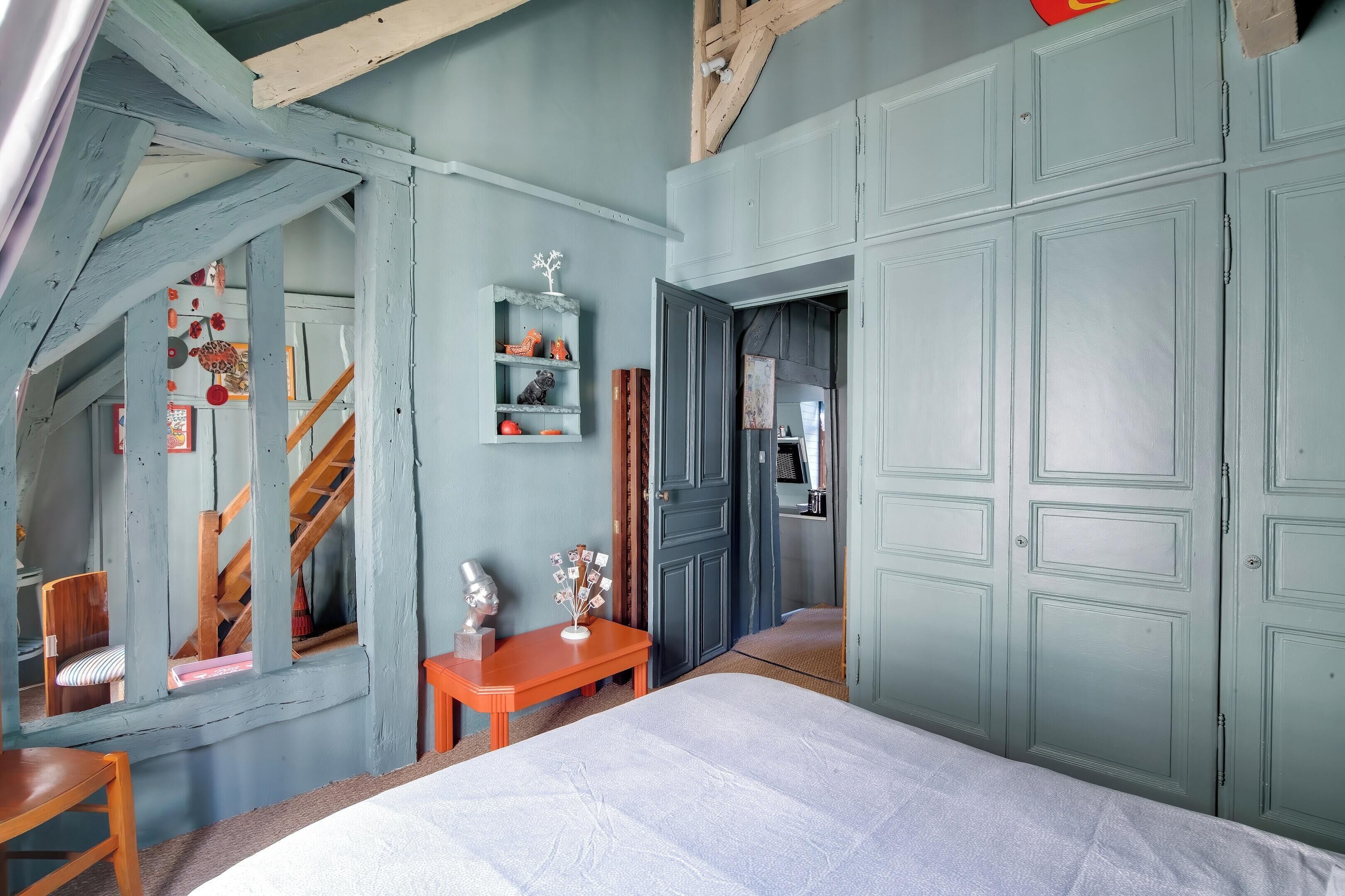 Quarto com cama, cadeira de madeira, pequena mesa, prateleira com objetos decorativos e escada para um andar superior.