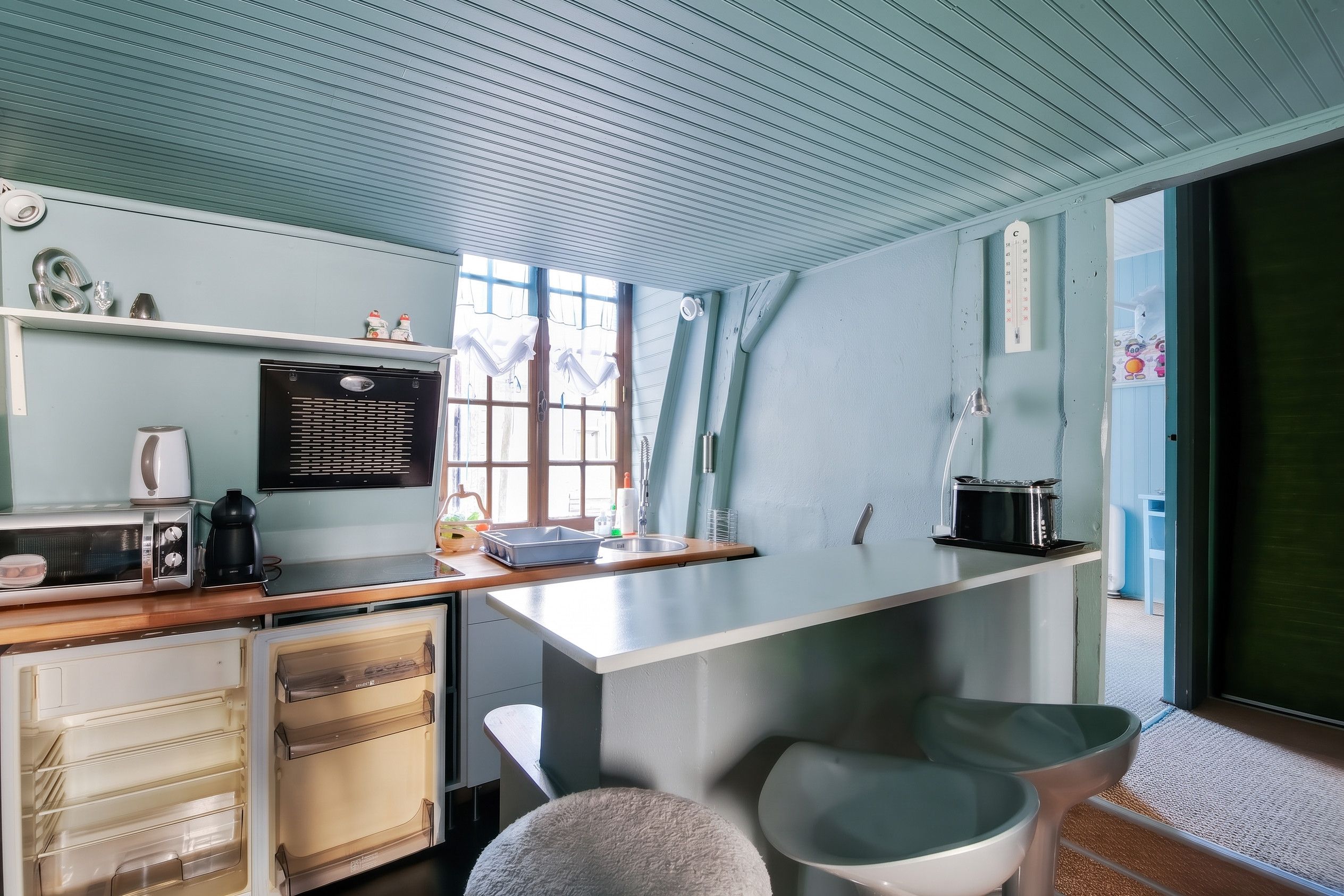 Eine kleine Küche mit Kühlschrank, Mikrowelle und Herd. Ein Fenster mit Vorhängen, ein Spülbecken und eine kleine Arbeitsfläche sind vorhanden.