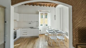 Una cocina con armarios blancos, una mesa de comedor con sillas y un ventanal con contraventanas.