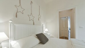 Un dormitorio con una cama blanca, una mesita de noche con lámpara y una puerta que da a otra habitación.