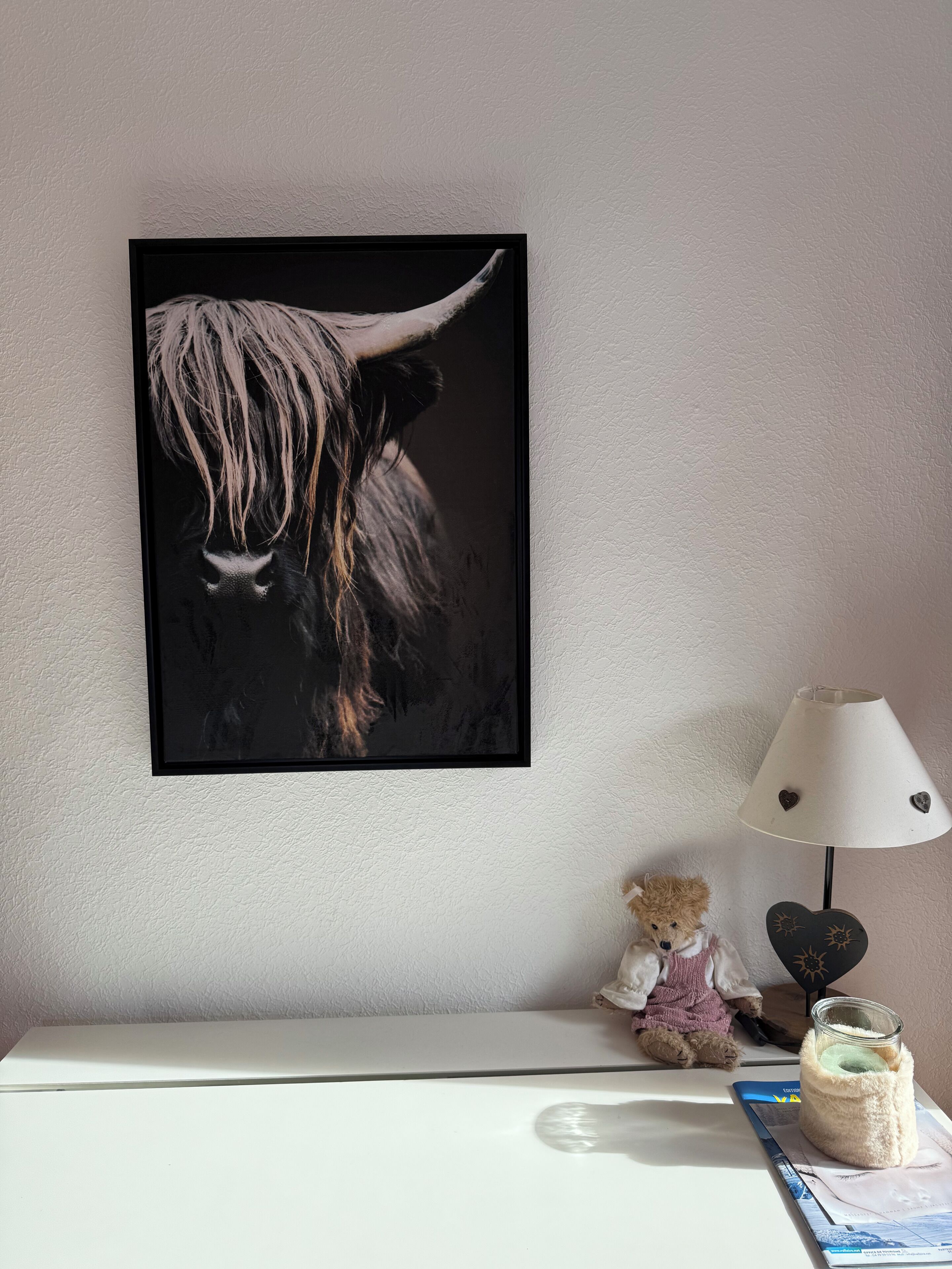 Una obra de arte en marco que muestra un yak, un osito de peluche pequeño, una lámpara y un frasco sobre una mesa.