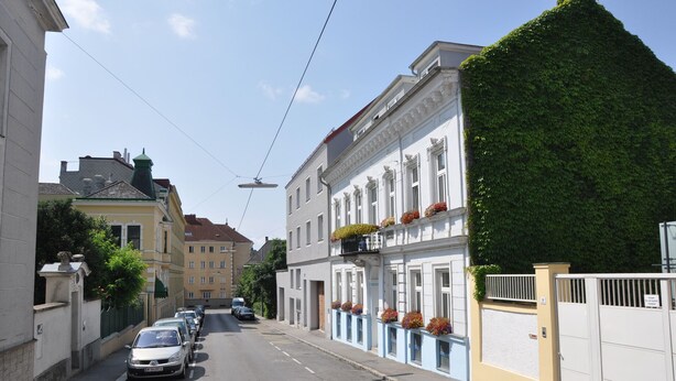 A street with parked cars, buildings with balconies, and a clear sky. - Livienna – Viennese period charm with garden, pool & fireplace near Schönbrunn (Wien)