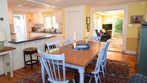 A dining area with a wooden table and white chairs, a kitchen in the background, and a view of the outdoors through a sliding door. - Spacious, Private Cape Cod Cottage with Central Air (Harwich Port)