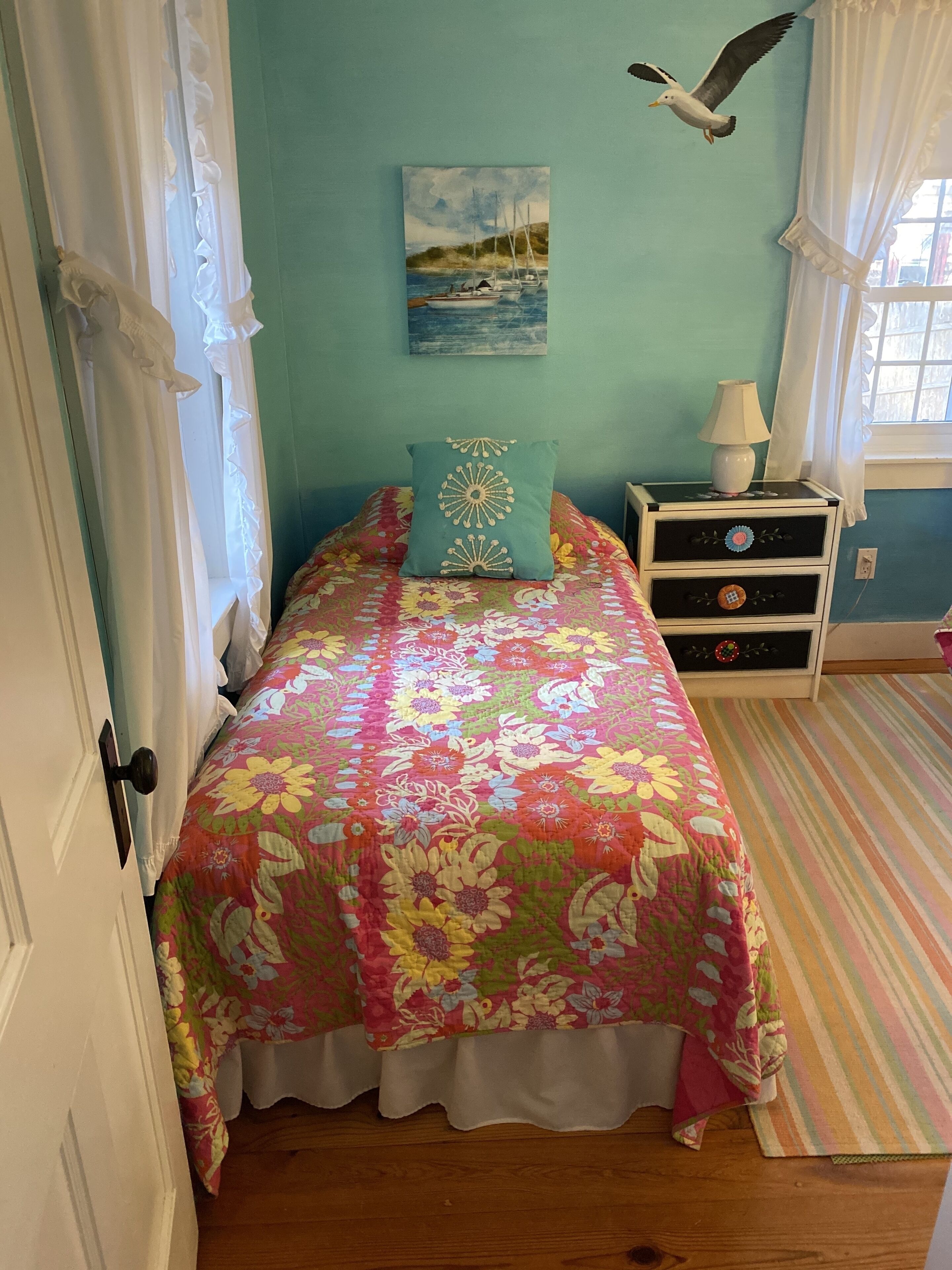 A bedroom with a floral bedspread, a teal pillow, a nightstand with a lamp, and a painting of a seascape.