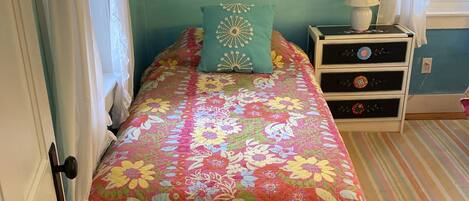 A bedroom with a floral bedspread, a teal pillow, a nightstand with a lamp, and a painting of a seascape.