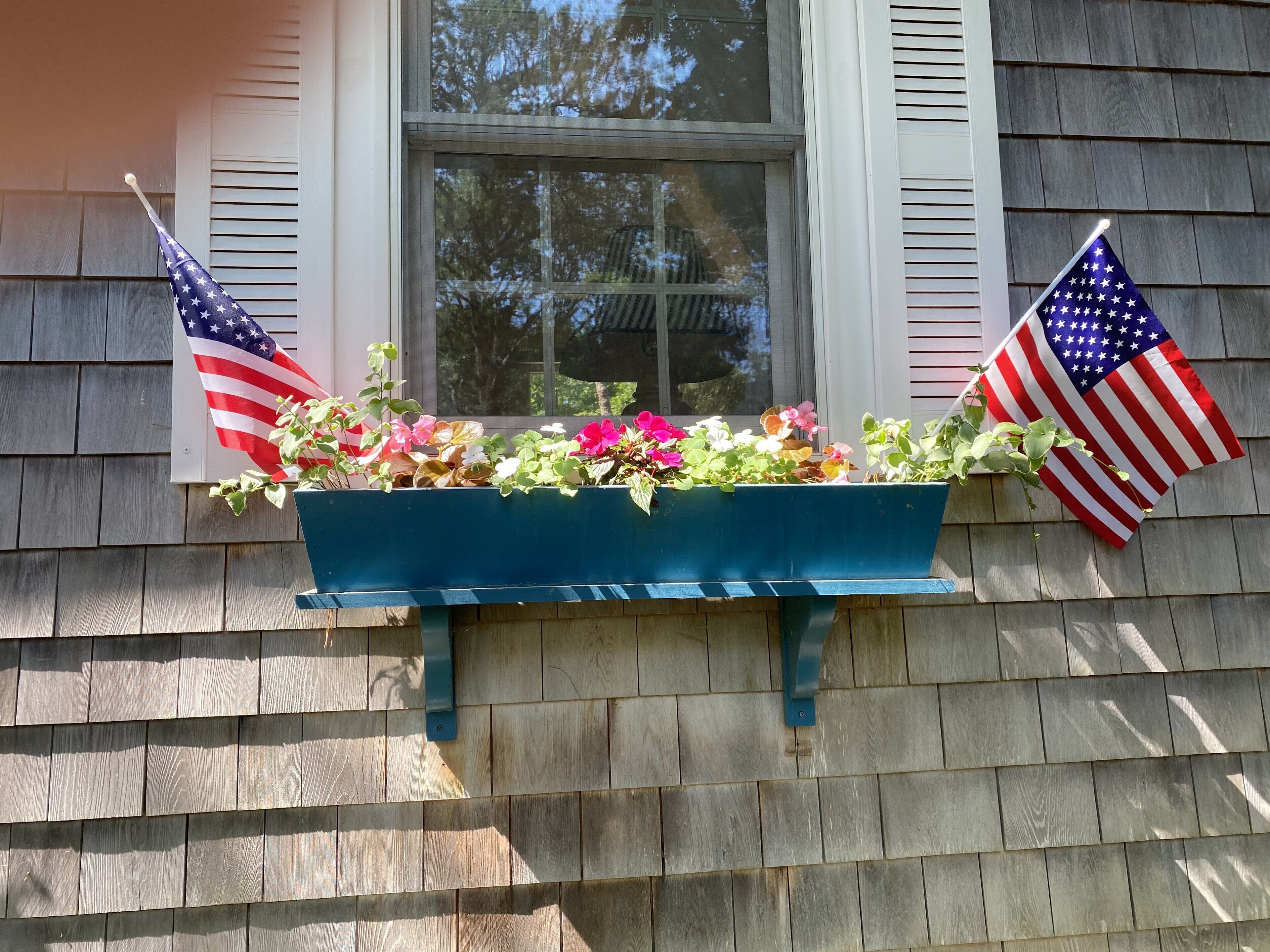 A window box with colorful flowers and two American flags on a house with wooden siding.