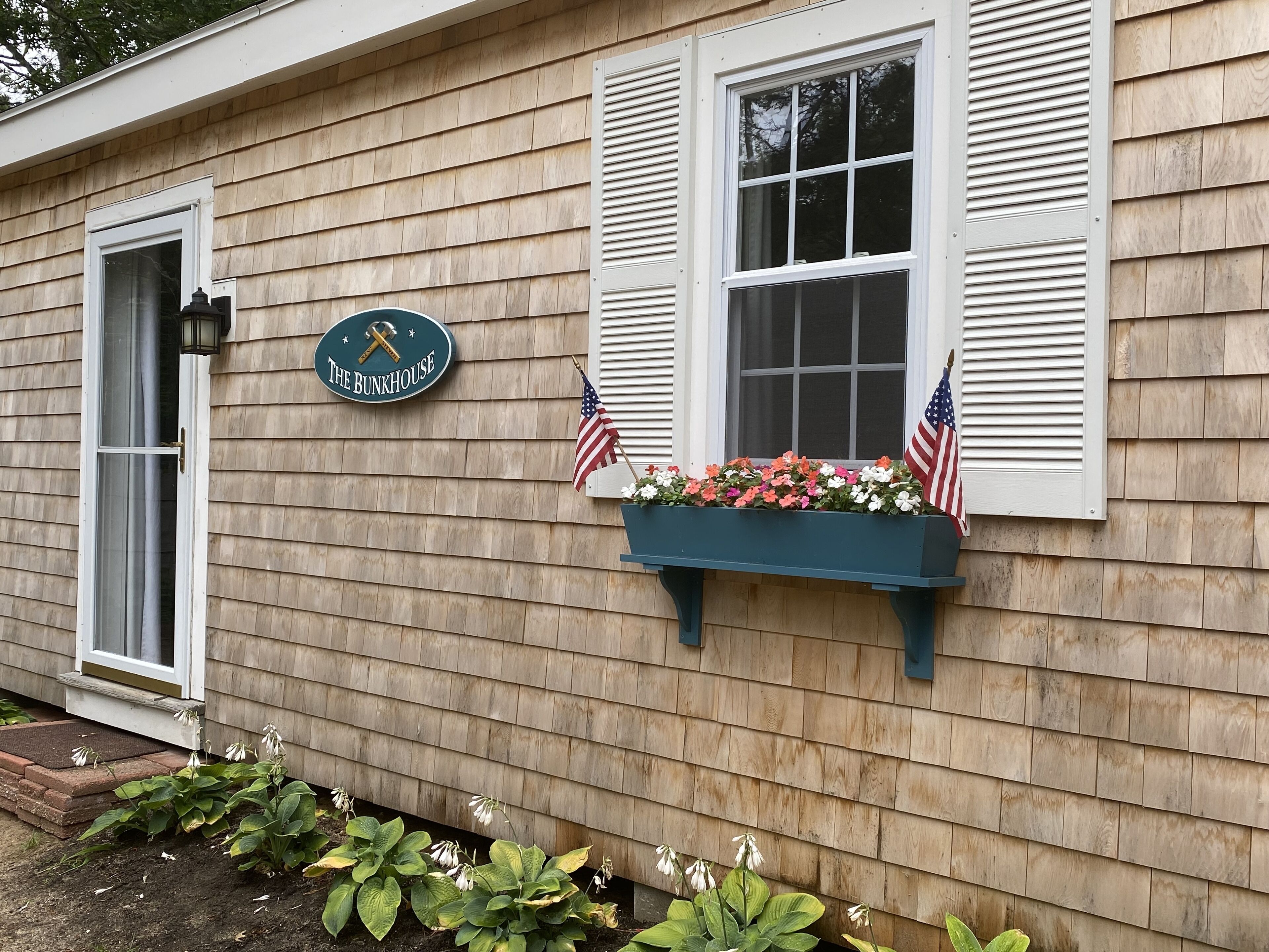 A house with a sign reading "The Bunkhouse," a window with an American flag, and a flower box with flowers.