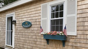 A house with a sign reading "The Bunkhouse," a window with an American flag, and a flower box with flowers. - Spacious, Private Cape Cod Cottage with Central Air (Harwich Port)