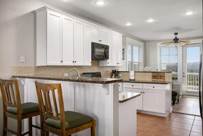 A kitchen with white cabinets, a microwave, a sink, and a bar area with two wooden chairs.