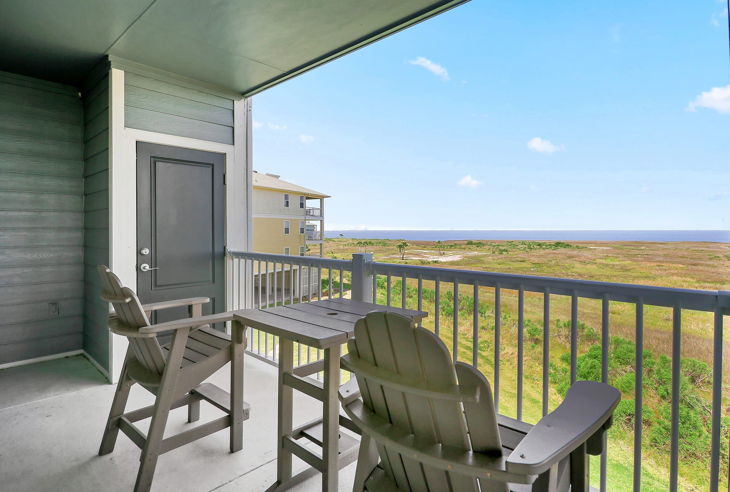 A balcony with two Adirondack chairs and a table, overlooking a grassy landscape and a building in the distance.