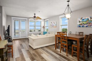Un vaste salon comprenant un canapé, une table à manger et des chaises, de grandes fenêtres équipées de stores et un ventilateur de plafond.