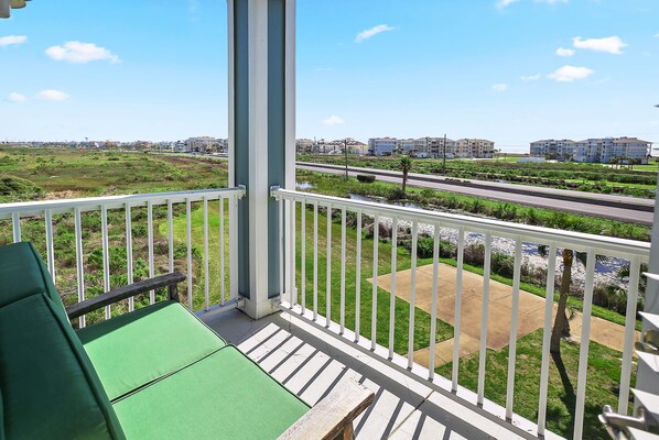A balcony with a green cushioned chair, a white railing, and a view of a road and buildings in the distance.