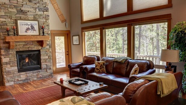 A cozy living room with a stone fireplace, leather sofas, a coffee table, and large windows offering a view of the outdoors. - Premier Luxury Home with 7 BR/7.5 BA, Hot Tub, Game Room, SHARC passes, A/C (Sunriver)
