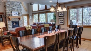 A spacious living room with a large wooden dining table, comfortable seating, a stone fireplace, and large windows offering a view of the outdoors. - Premier Luxury Home with 7 BR/7.5 BA, Hot Tub, Game Room, SHARC passes, A/C (Sunriver)