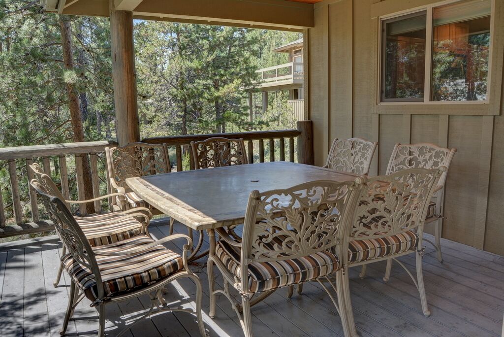 A covered outdoor deck with a wooden table and six chairs, surrounded by a wooden railing.