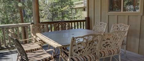 A covered outdoor deck with a wooden table and six chairs, surrounded by a wooden railing.