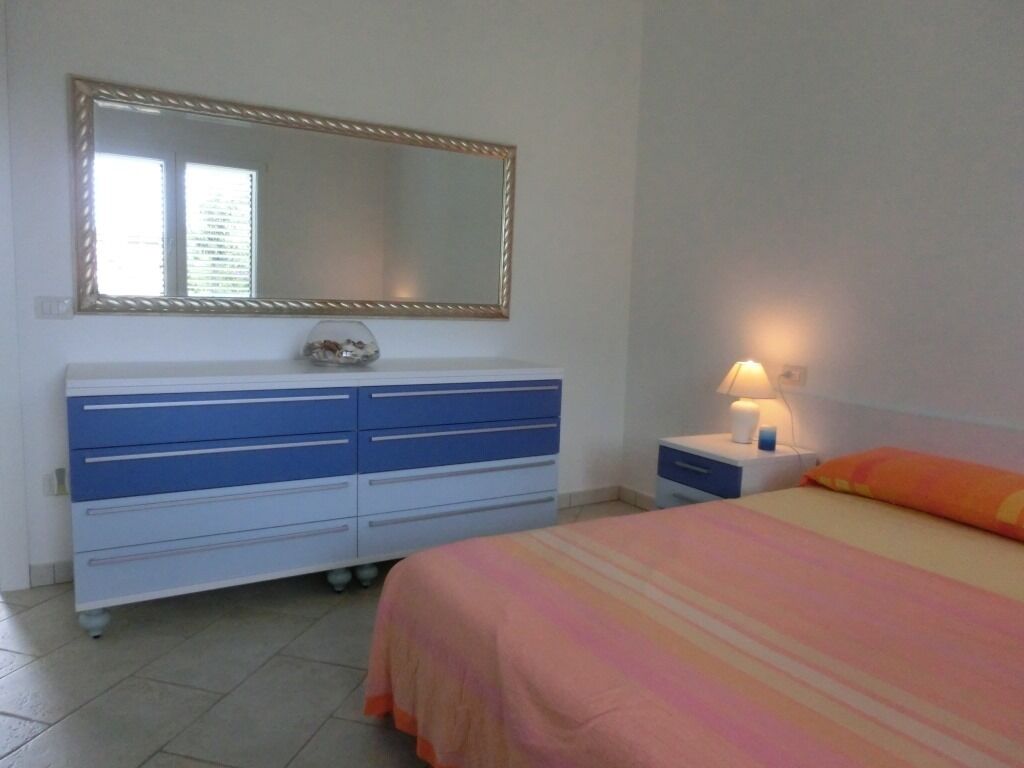 A bedroom with a bed, a dresser with drawers, a mirror, a nightstand with a lamp, and a window with blinds.