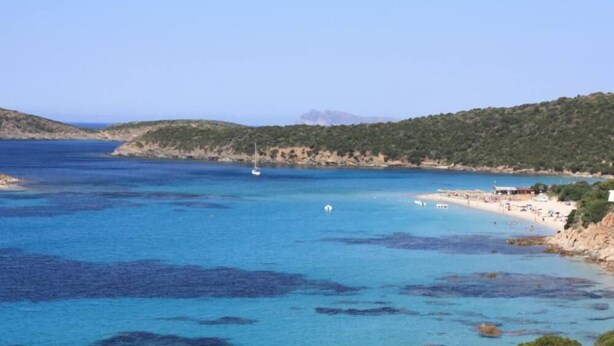 Une vue sur la côte, avec une eau bleue et limpide, une plage de sable et un paysage vallonné en arrière-plan.