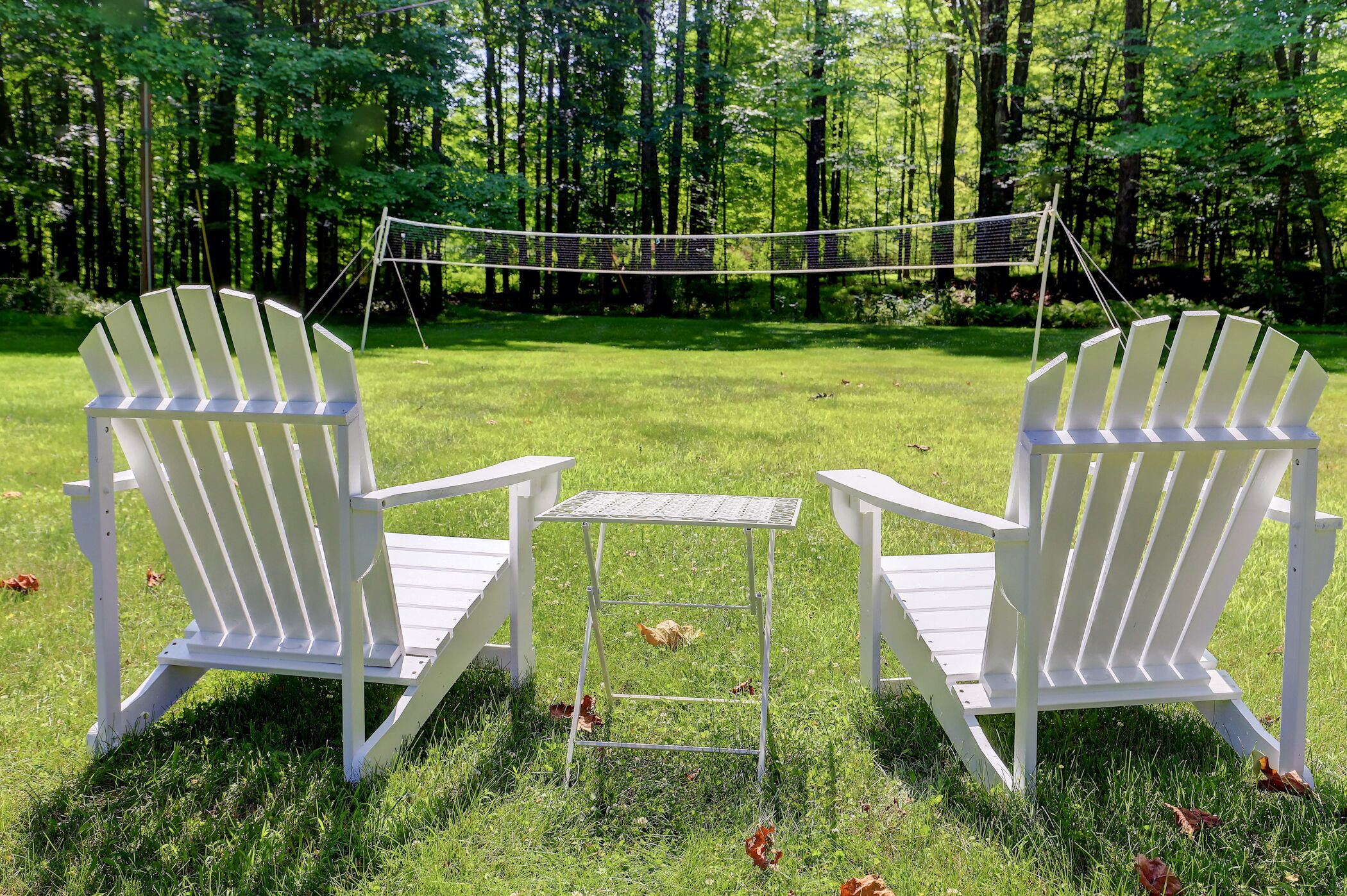 Deux chaises Adirondack blanches, accompagnées d’une table en métal, sont placées dans un espace extérieur verdoyant, avec des arbres en arrière-plan.