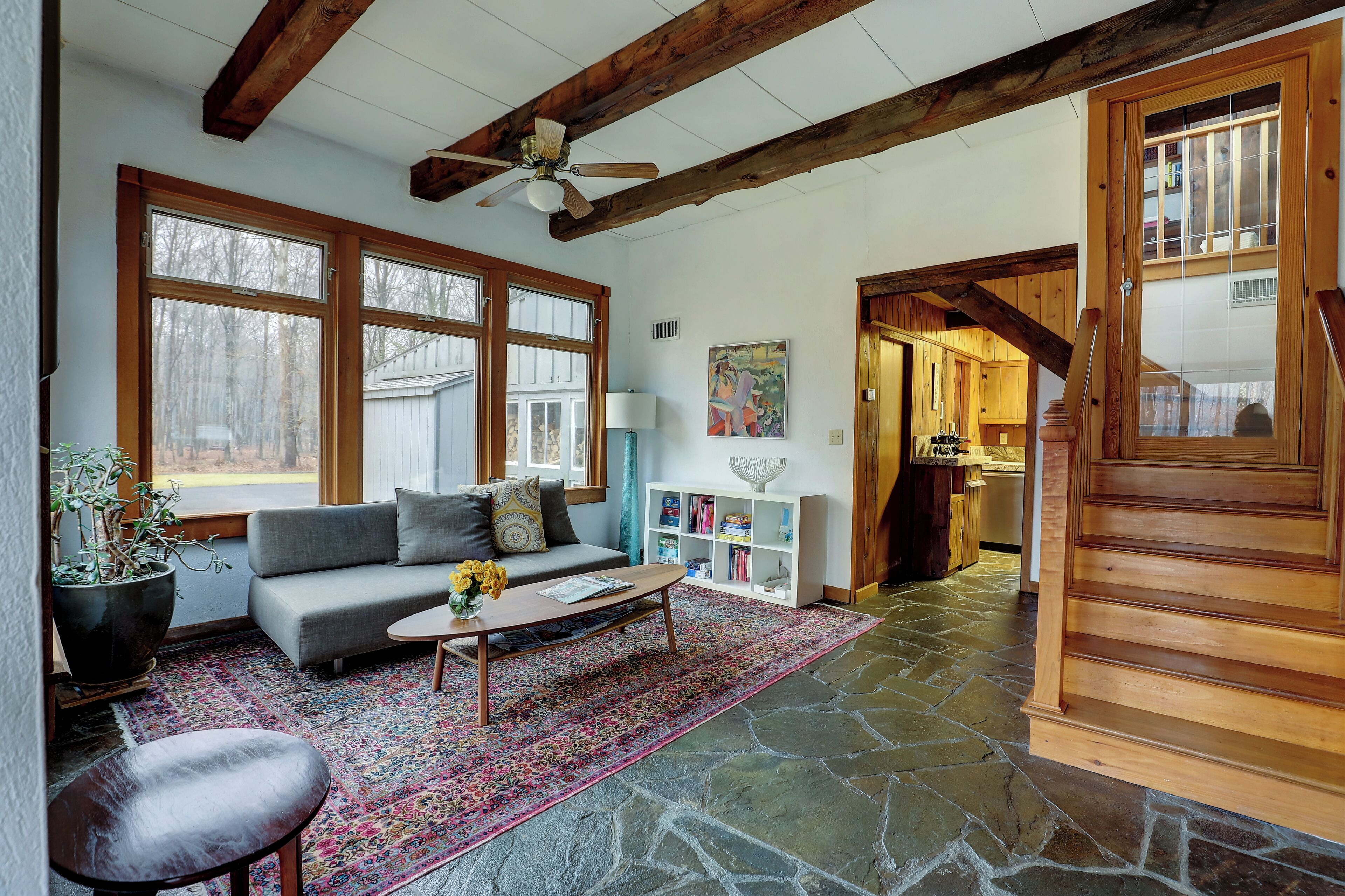 A spacious living room with a stone floor, a gray sofa, a coffee table, and a large window with a view of trees.