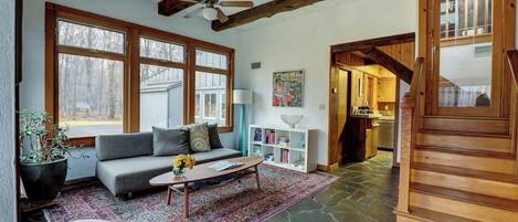 A spacious living room with a stone floor, a gray sofa, a coffee table, and a large window with a view of trees.