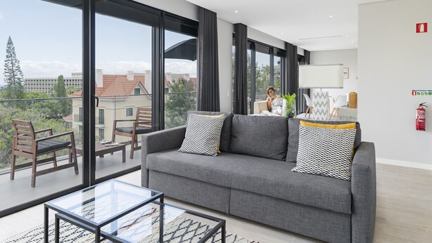 Una sala de estar moderna con un sofá gris, dos sillones, una mesa de centro de vidrio y amplias ventanas con vistas a edificios y árboles.