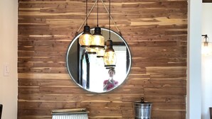 Eine Holzwand mit einem runden Spiegel, ein schwarzer Tisch mit einem Metallbehälter und eine Pendelleuchte.