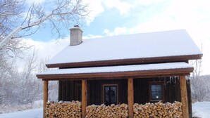 Eine Holzhütte mit verschneitem Dach und Schornstein, umgeben von Holzstapeln.