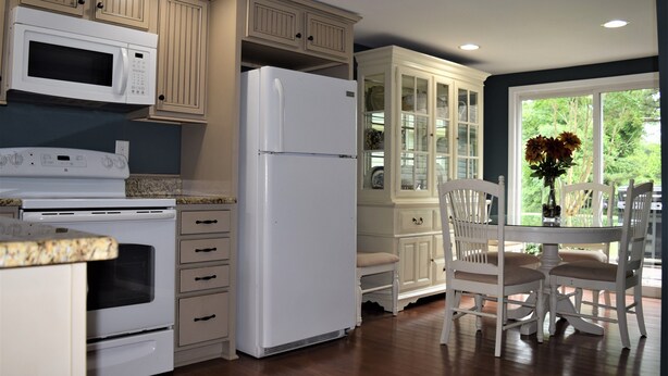A kitchen with a white refrigerator, stove, and microwave, wooden cabinets, and a dining area with a table and chairs. - Moonshine Cottage - Time to GOLF! (Pinehurst)