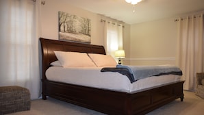 A neatly made bed with a wooden headboard, a bedside lamp, and a chair in a well-lit bedroom with a large window and a painting on the wall. - Moonshine Cottage - Time to GOLF! (Pinehurst)