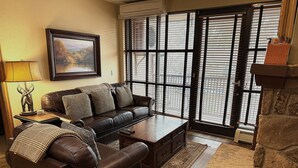 A living room with a brown leather sofa, a coffee table, a rug, a framed picture on the wall, and a large window with blinds. - In the heart of Avon -walk to restaurants & gondola!  Immaculate 2bed/2bath (Avon)