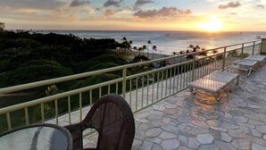 Une terrasse avec un sol en pierre, une rambarde en métal et une vue sur l’océan au coucher du soleil.