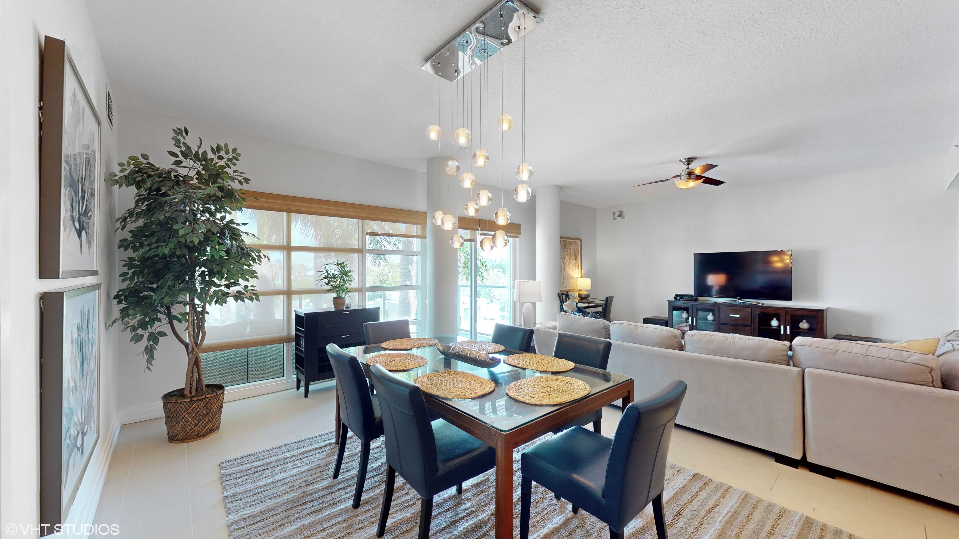 Una sala de estar moderna con un televisor grande, una zona de comedor con una mesa de vidrio y un ventilador de techo.