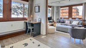 A modern living room with a wooden desk, a grey sofa, and a view of a snowy mountain. - Renovated apartment 64m², 6 pers, balcony, Wi-Fi, slopes (La Plagne)