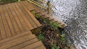 A wooden deck with a railing leading to a calm body of water surrounded by greenery.