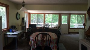 A cozy kitchen with a dining table, chairs, and a fan. There are large windows providing natural light and views of the outdoors.