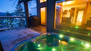 A hot tub with green lights on a wooden deck with a view of a house and snow-covered trees.
