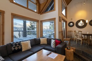 A cozy living room with a sectional sofa, wooden coffee table, and a view of snowy mountains through large windows. - TRUE Ski inSki out Hot Tub on Village Run-TRULY Slopeside-Luxury, Amazing Views. (Whistler)