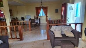 Un grand espace de vie comprenant un canapé, une table basse et une table à manger avec des chaises. Au fond, on aperçoit un coin cuisine et une porte donnant sur l’extérieur.