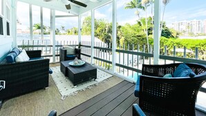 Un espacio al aire libre cubierto con mobiliario de mimbre, un ventilador de techo y vistas a palmeras y edificios.