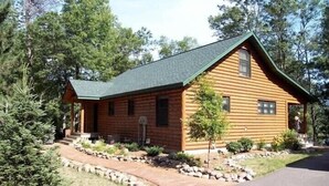 A log cabin with a green roof, a stone pathway leading to the entrance, and a small tree in front. - Luxury Lakefront Retreat on a Quiet Lake (Spooner)