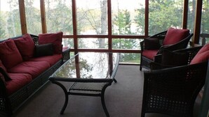 A room with a red sofa and two wicker chairs, a glass coffee table, and large windows offering a view of trees. - Luxury Lakefront Retreat on a Quiet Lake (Spooner)