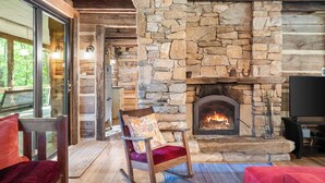 A cozy room with a stone fireplace, wooden beams, and a rocking chair with a colorful cushion. - Restored Double Log Cabin on 400 Mountain Acres (Covesville)