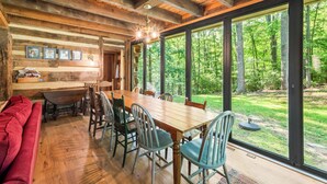 A rustic wooden interior with a large dining table, chairs, and a red sofa. Large windows offer a view of a lush green forest. - Restored Double Log Cabin on 400 Mountain Acres (Covesville)