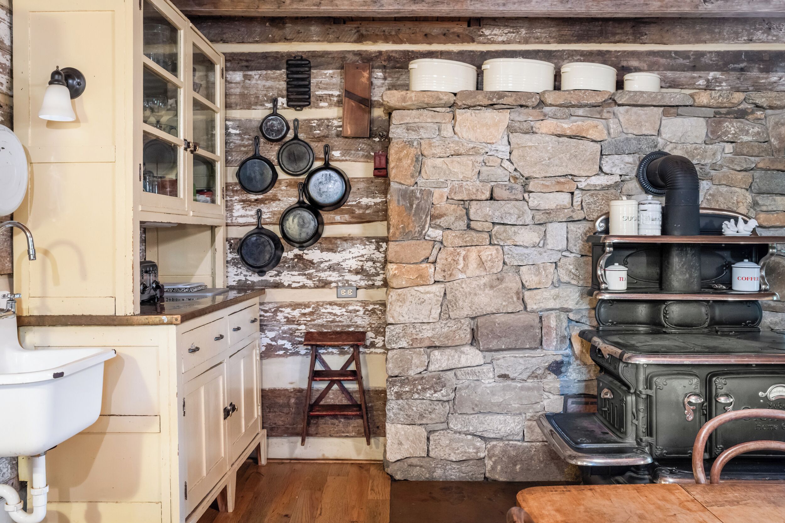 Stylish rustic kitchen with cream cabinetry, farmhouse sink, vintage décor, and a striking stone wall with a classic wood-fired stove—where charm meets functionality