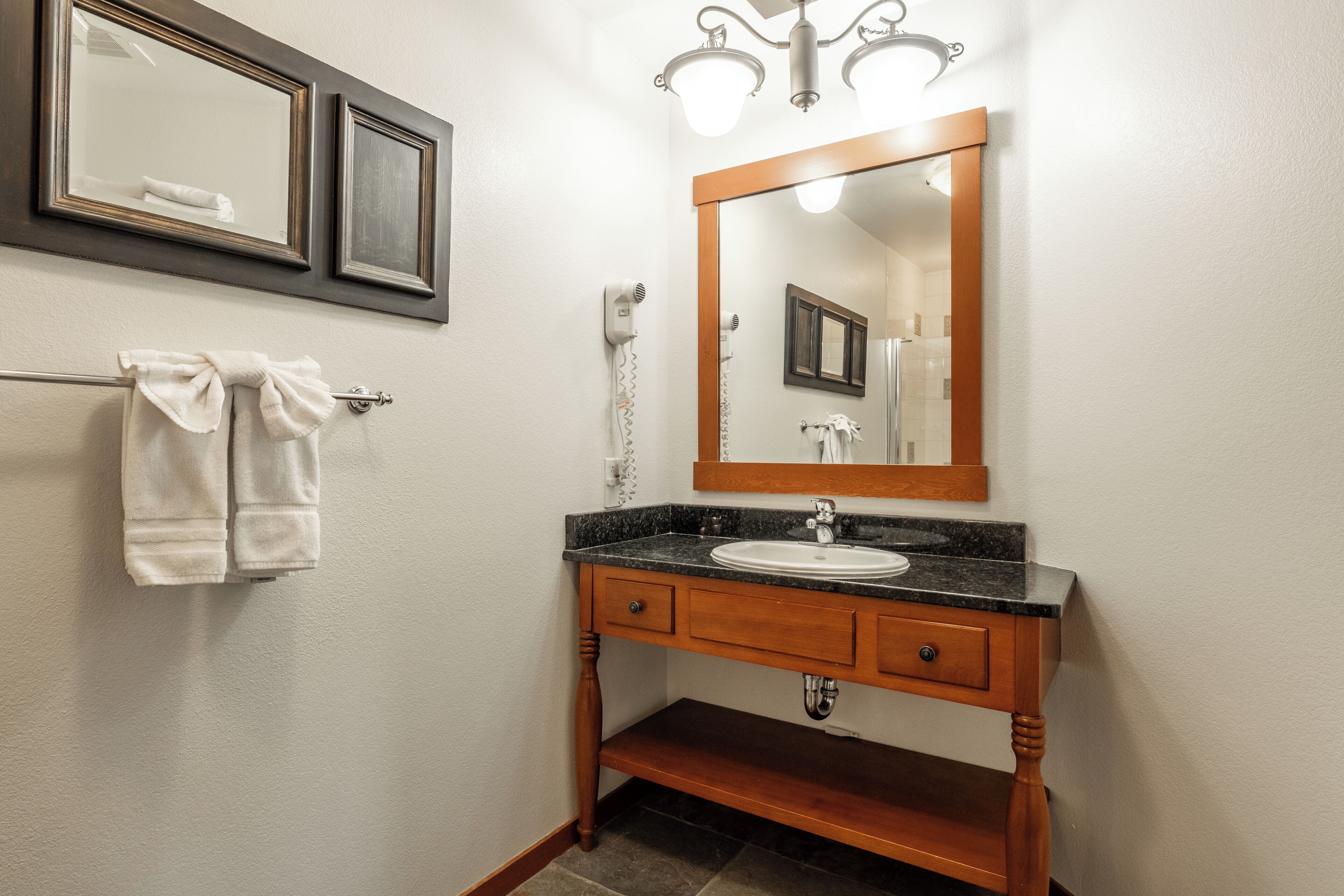 A bathroom with a wooden vanity, a sink, and a mirror. There are towels hanging on a rack and a hairdryer on the wall.