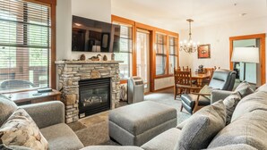 A living room with a stone fireplace, grey sofas, a dining area with a table and chairs, and large windows with blinds.