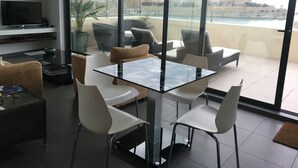 Ein modernes Wohnzimmer mit Glastisch, weißen Stühlen, einer Couch und einem Fernseher. Durch große Fenster ist ein Balkon mit Sitzgelegenheit und Blick auf die Stadt zu sehen.